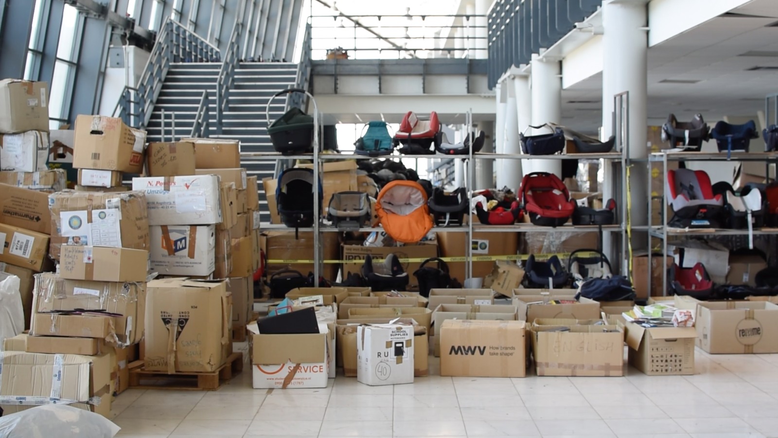 Lots of boxes with donations stacked on the floor next to a shelf with car seats for children.